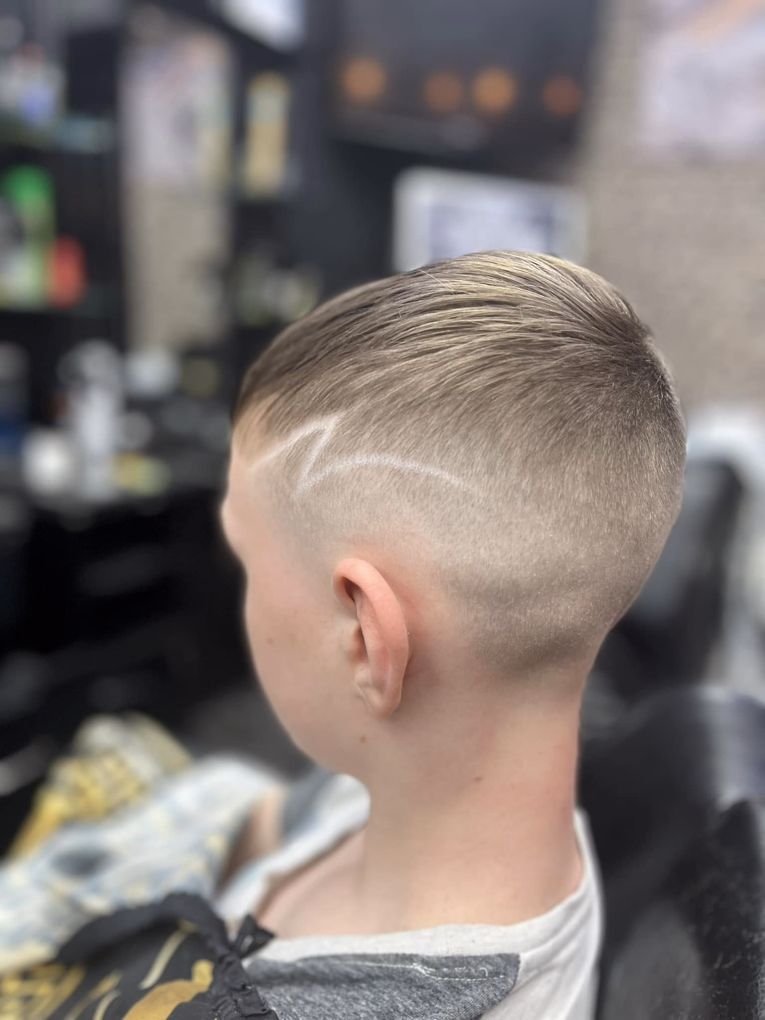 High skin fade with a curved design line shaved above the ear and a fine blonde top swept back, rear three-quarter view